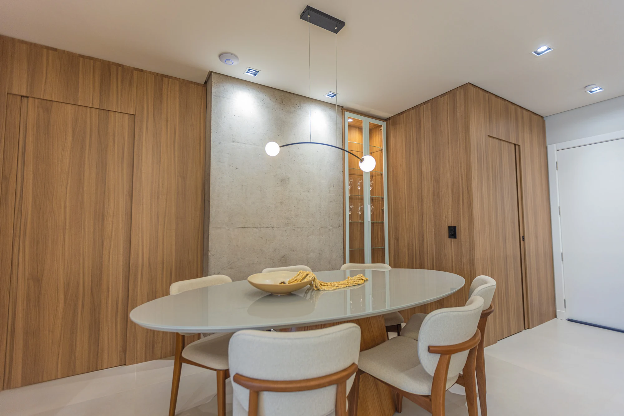 Mesa de jantar clara com cadeiras estofadas, painel de madeira e pilar de concreto aparente